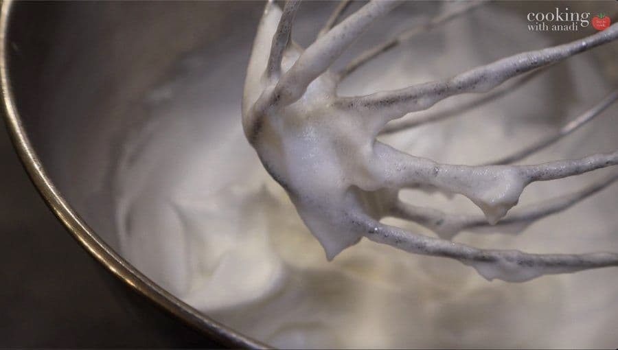 Whipped egg whites with soft peaks forming on mixer whisk over bowl.