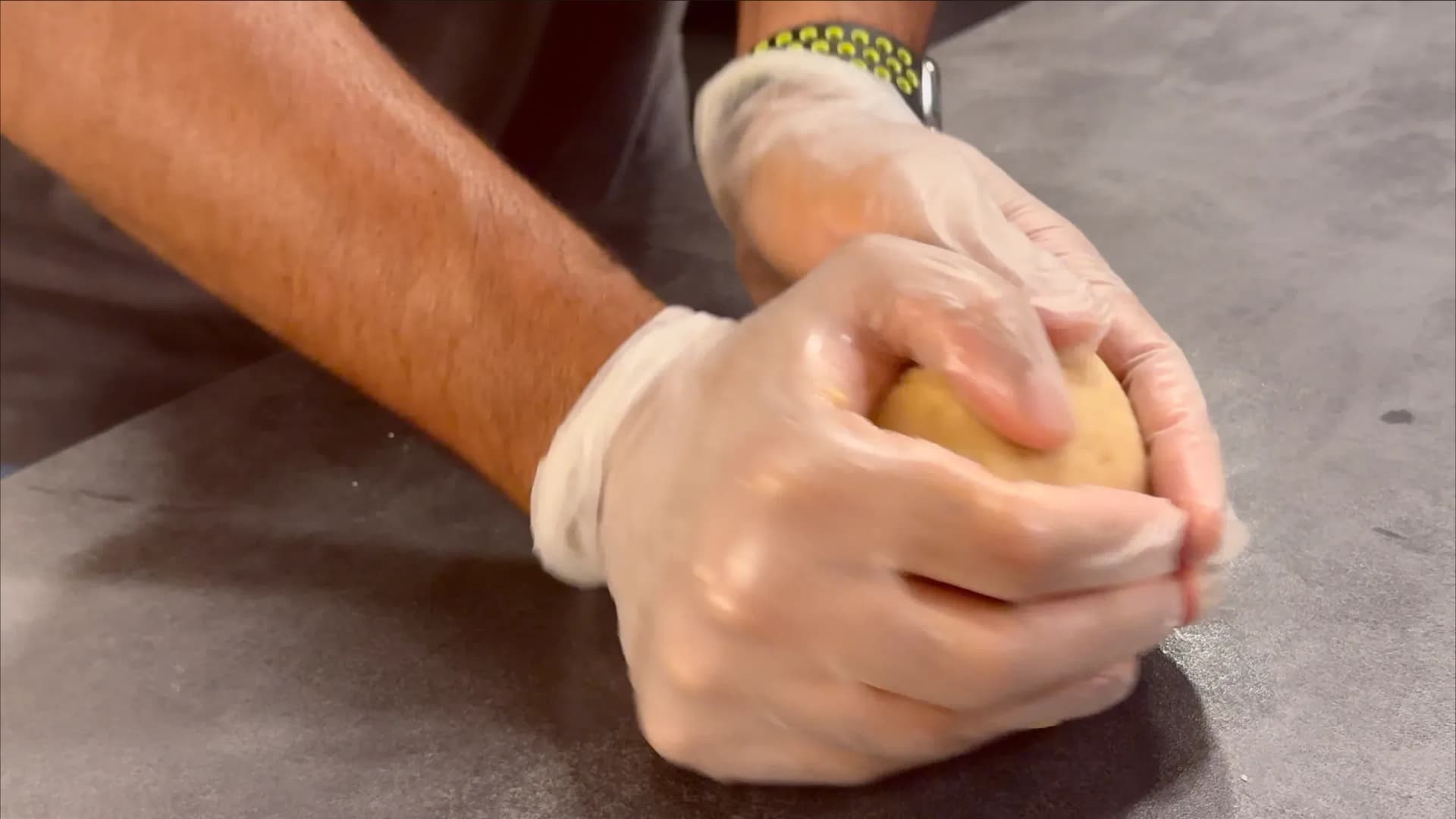 Hands in clear plastic gloves forming pastry dough into a ball on a dark surface.