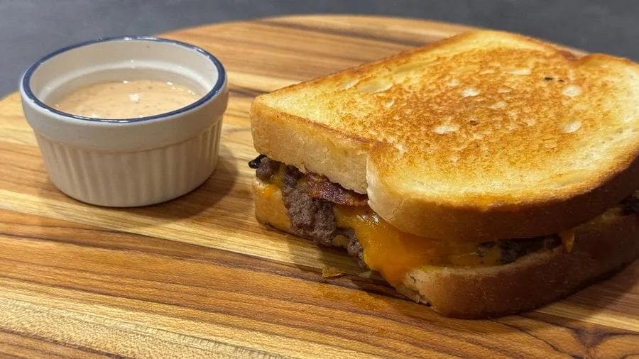 Golden-brown patty melt with melted cheese and bacon on toasted bread, served with a side of sauce on a wooden board.