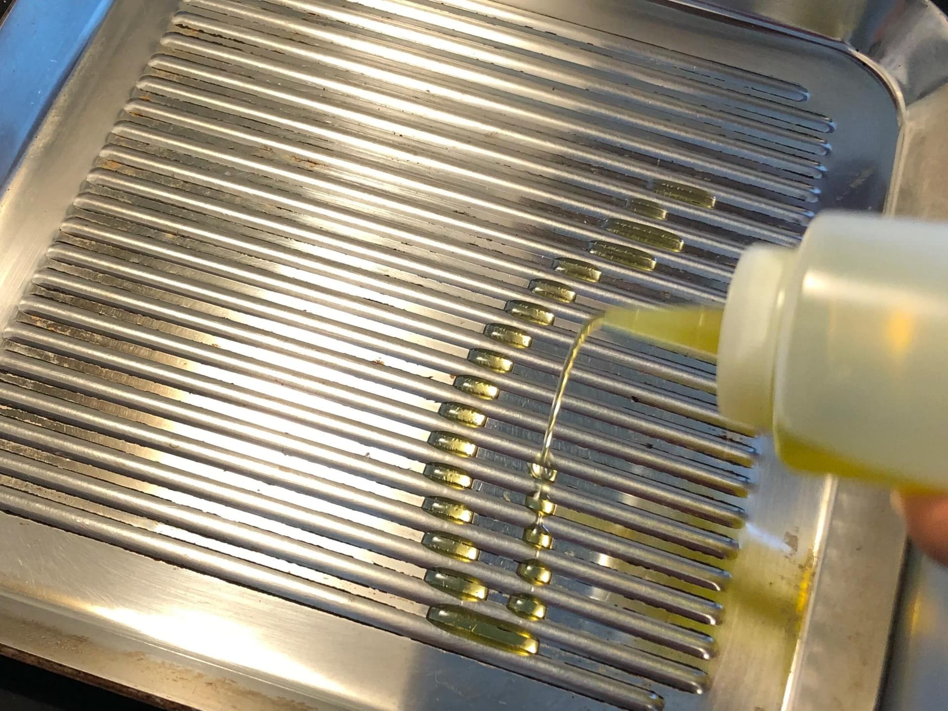 Multiple metal drill bits arranged in a metal tray with oil being applied from a bottle