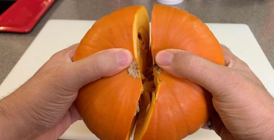 Hands pulling apart a halved pumpkin to show the interior flesh and seeds.