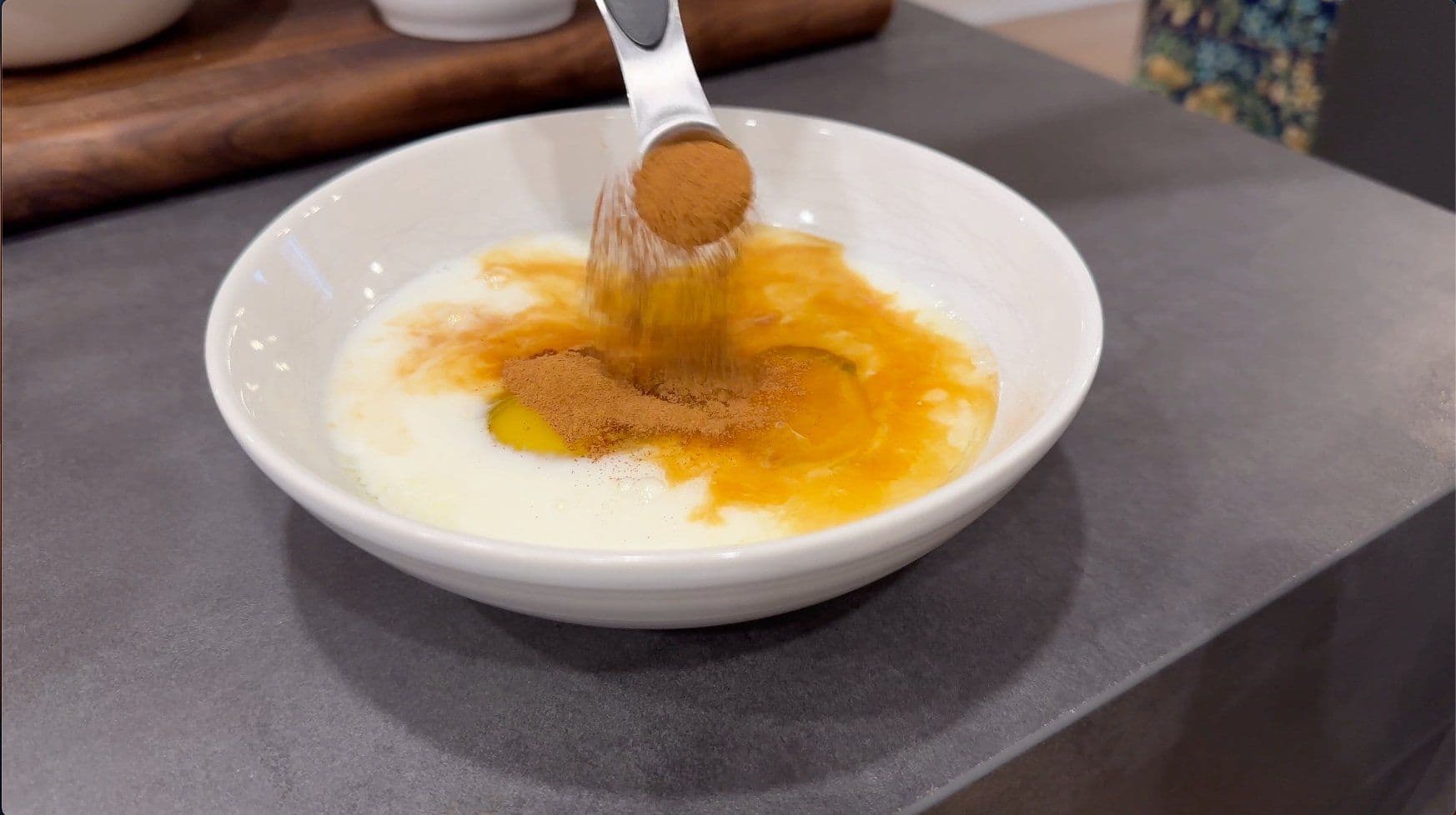 Cinnamon and nutmeg being sprinkled over cream-colored egg mixture in a white bowl.