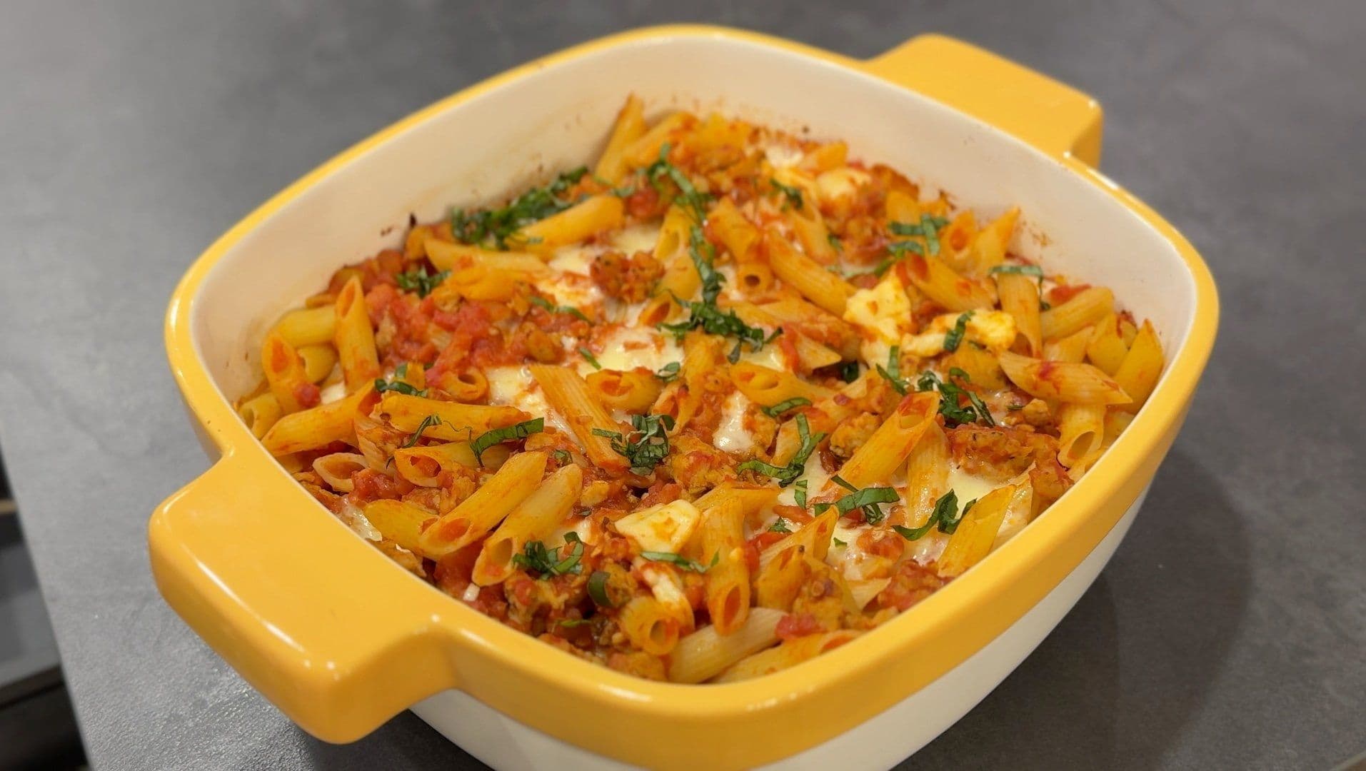Cheesy baked penne with Italian sausage in a yellow casserole dish, topped with melted cheese and fresh basil.