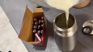Cream being poured into a metal whipped cream dispenser next to an open box of charger cartridges.