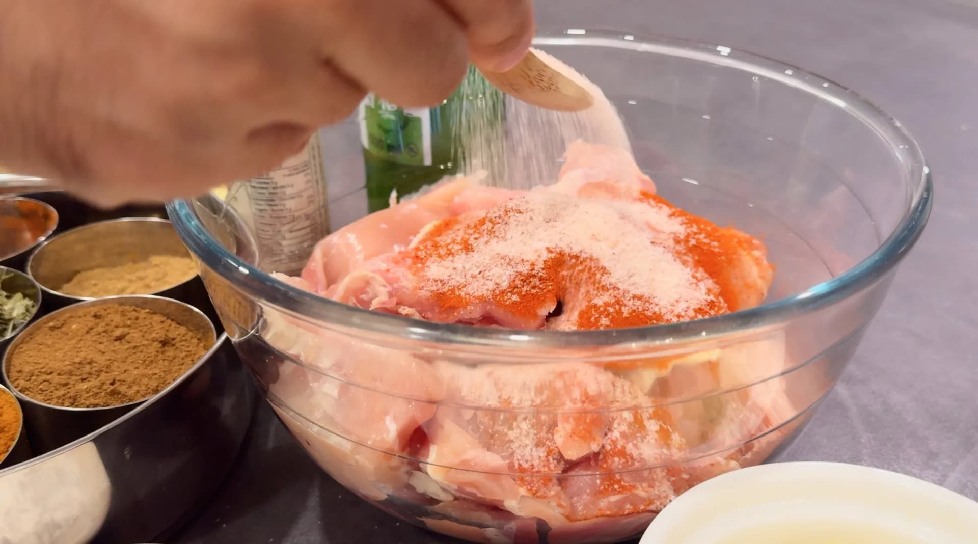 Salt being sprinkled over raw chicken thighs coated in red Kashmiri chili powder in a glass bowl.