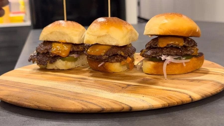 Three smash burger sliders with melted cheese on wooden skewers, served on a wooden board.
