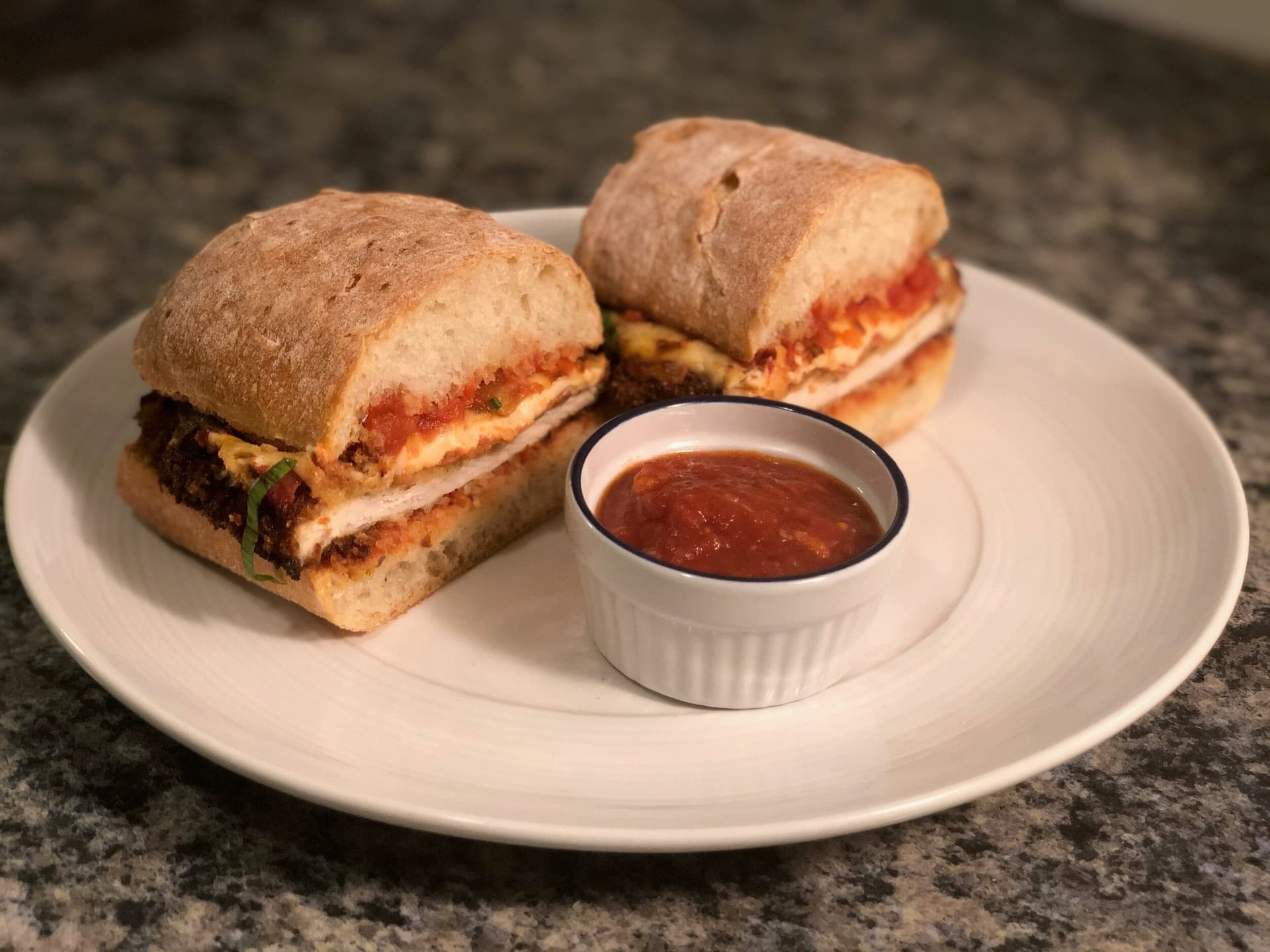 Two crispy chicken parm sandwiches on a white plate with a small bowl of marinara sauce for dipping.