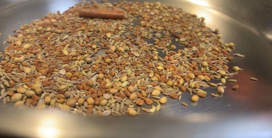 Toasted spice blend in a stainless steel pan, including cinnamon stick, cumin seeds, and coriander seeds, lightly browned and fragrant.