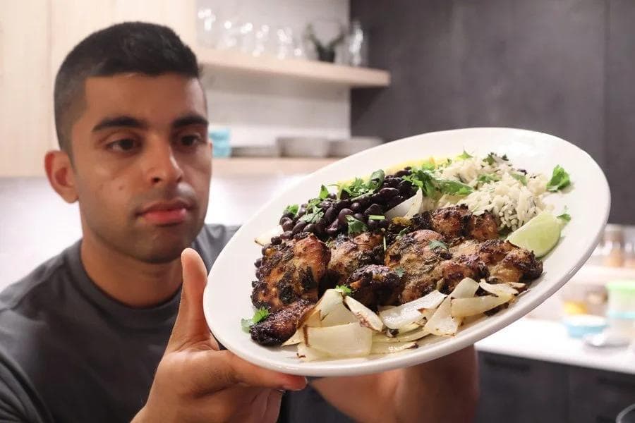 Man holding white plate of Cuban mojo chicken with black beans, rice, and lime wedges in kitchen.