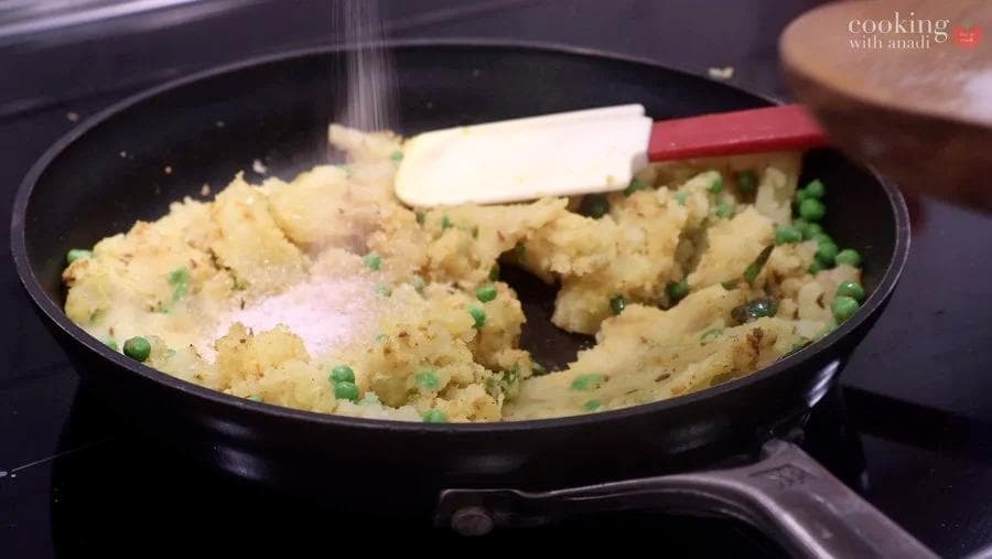 Butter being stirred into cooked potatoes and peas in a skillet during preparation of aloo matar curry.