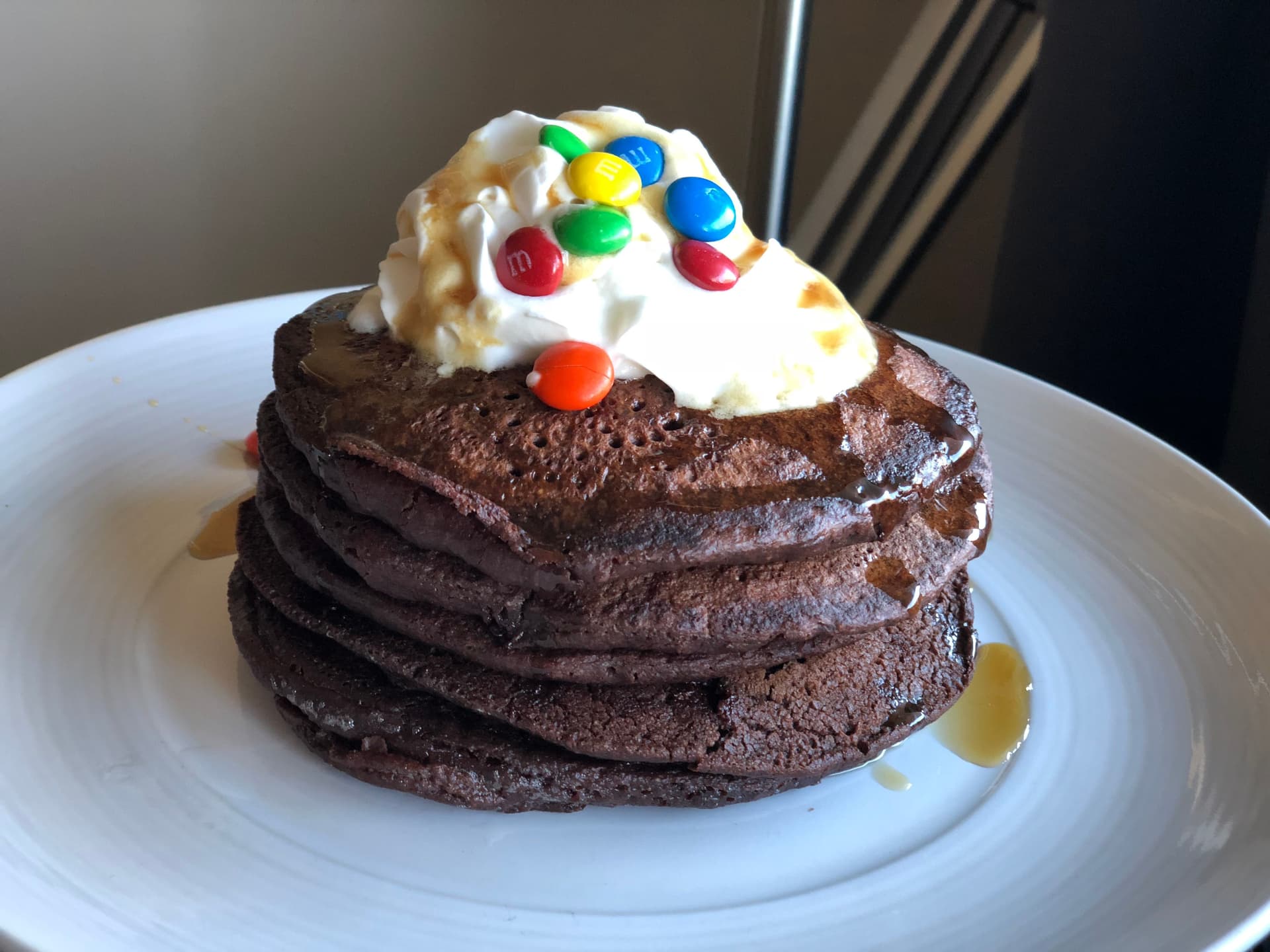 Stack of chocolate pancakes topped with whipped cream and colorful M&M candies on a white plate.