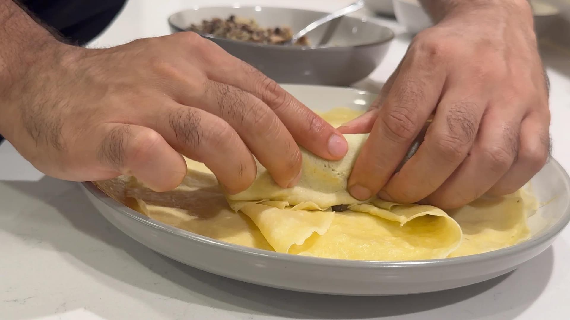 Hands folding a crepe with mushroom-sauerkraut filling on a white plate.