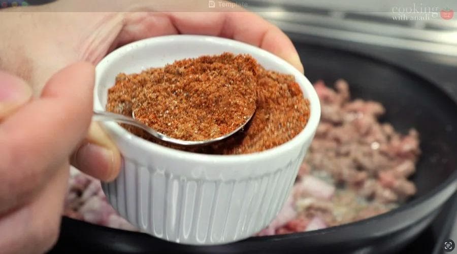 Taco seasoning being measured in a small white bowl with a spoon over a pan of browning ground beef.
