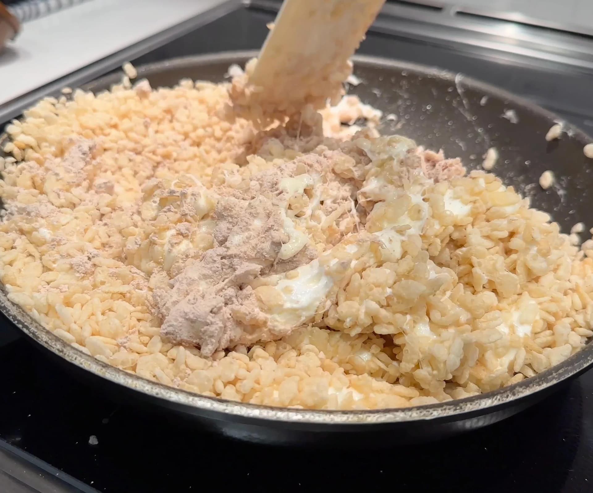 Wooden spoon stirring protein powder into rice cereal mixture in a dark pan on stovetop.