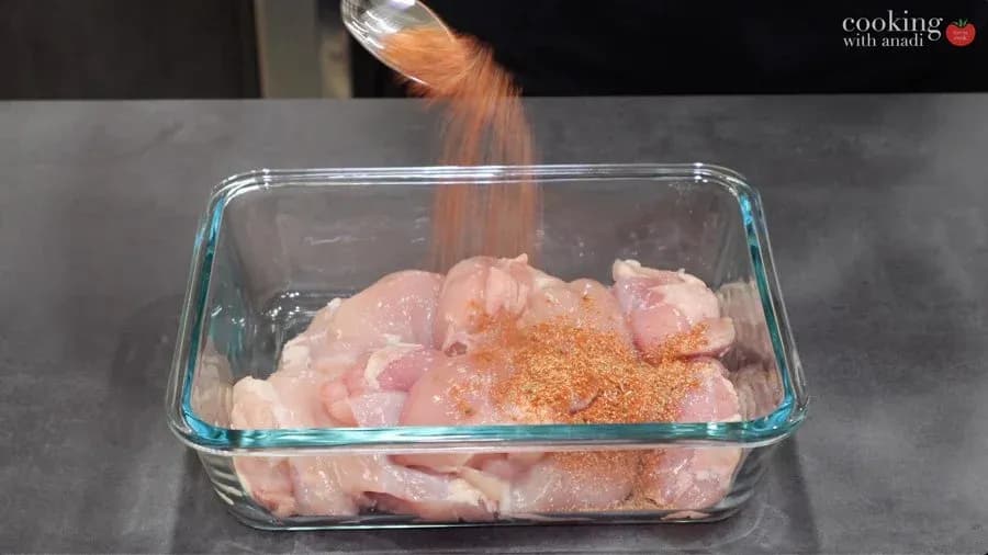 Spice mix being sprinkled over raw chicken thighs in a glass baking dish for marinating.