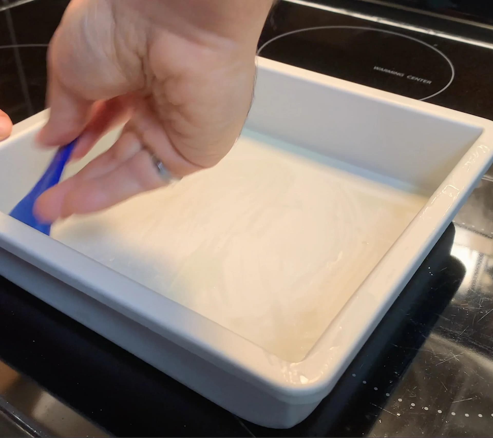 Hand pressing parchment paper into a greased white rectangular baking pan on a stovetop.