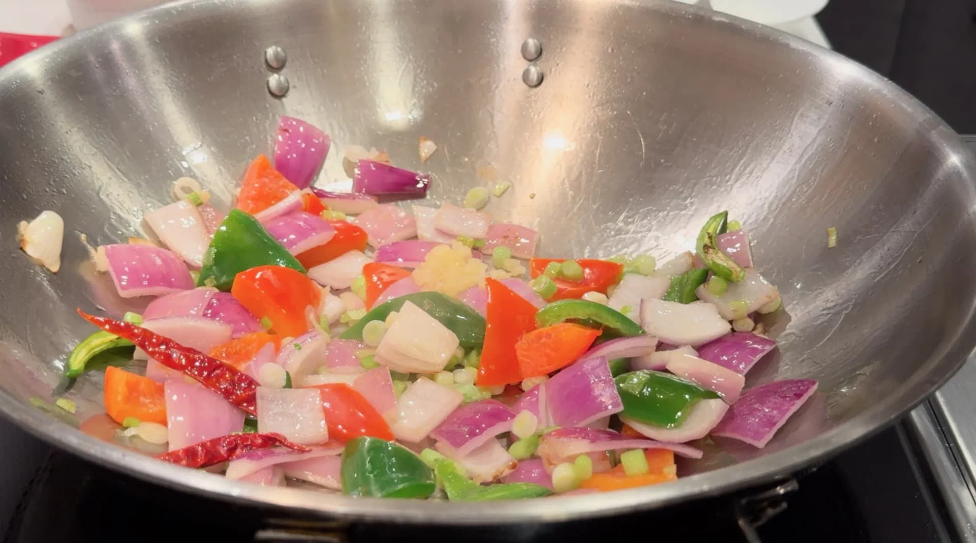 Chopped red onion, green and red bell peppers, dried chili, and green onion stir-frying in a steel wok.