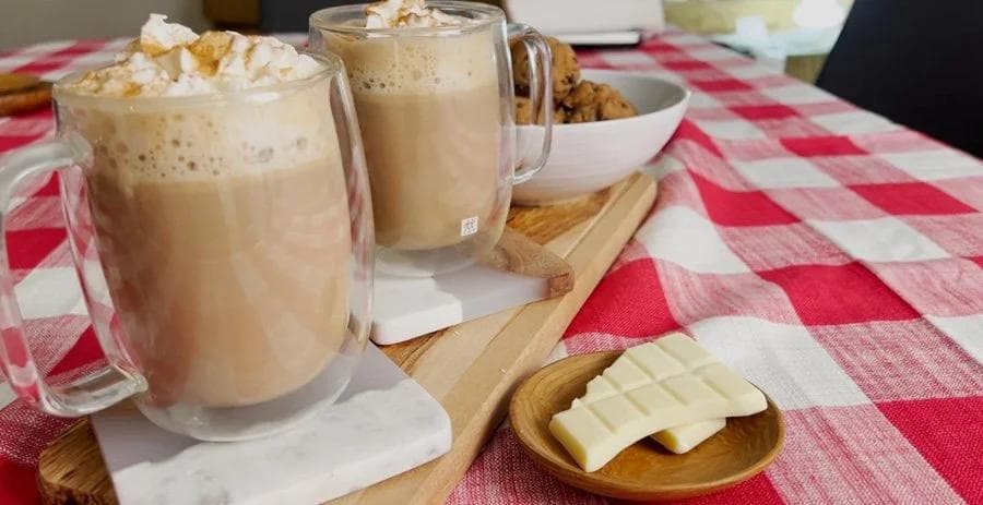 Two glasses of white chocolate mocha with whipped cream and toppings on a red gingham tablecloth with white chocolate squares and ingredients.