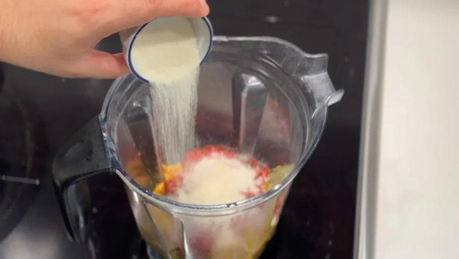 Pouring coconut milk into a blender containing peanut sauce ingredients including chili, garlic, and spices.