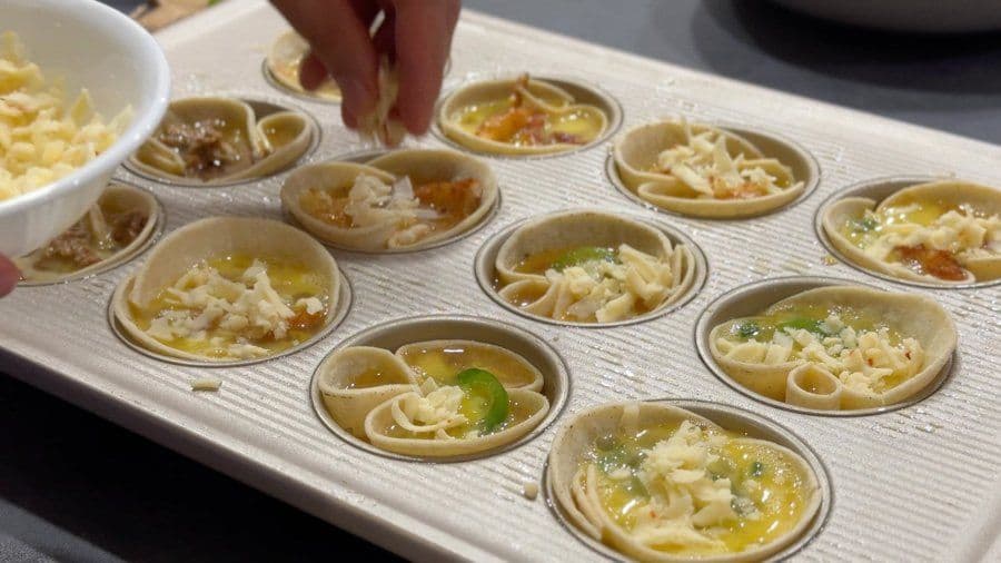 Baked egg cups in a muffin tin with pasta, cheese, and toppings being filled from a white bowl.