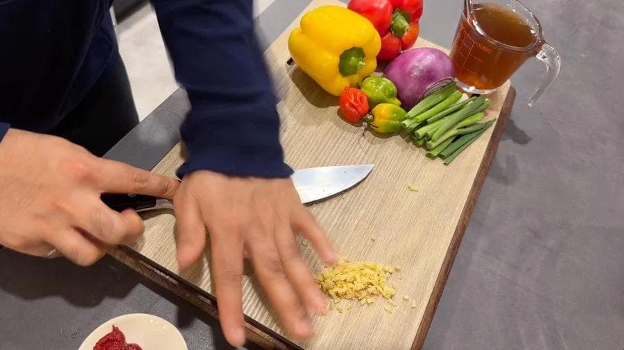 Hands chopping minced garlic on a wooden cutting board with fresh vegetables and a glass of broth nearby.