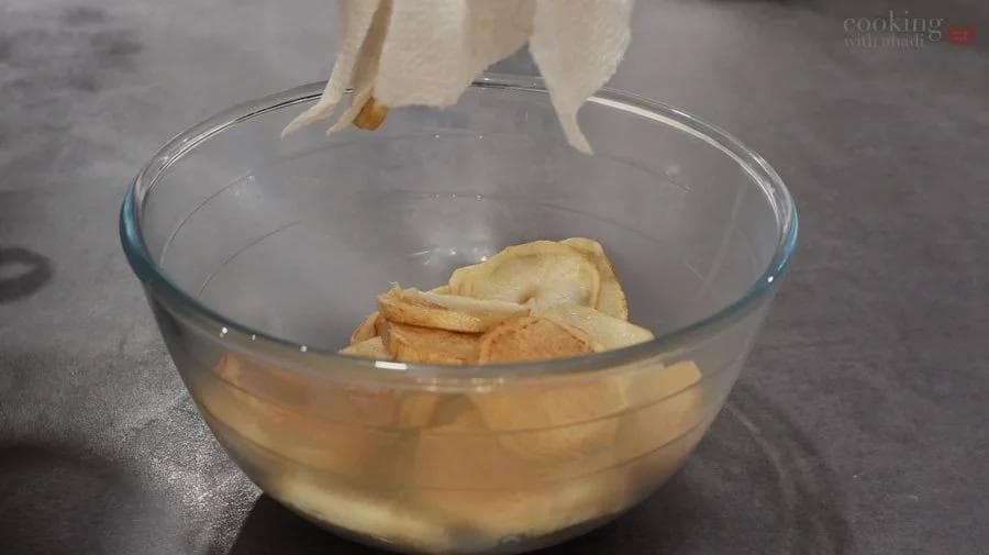 Sliced potatoes being removed from a glass bowl of golden oil after shallow frying for perkedel.