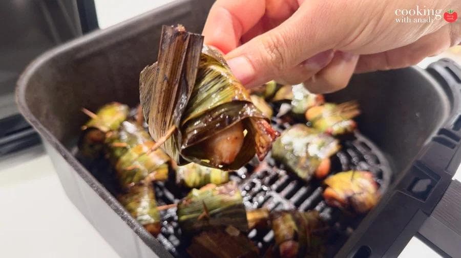 Hand holding a cooked pandan-wrapped chicken parcel over an air fryer basket filled with more golden-charred bundles.