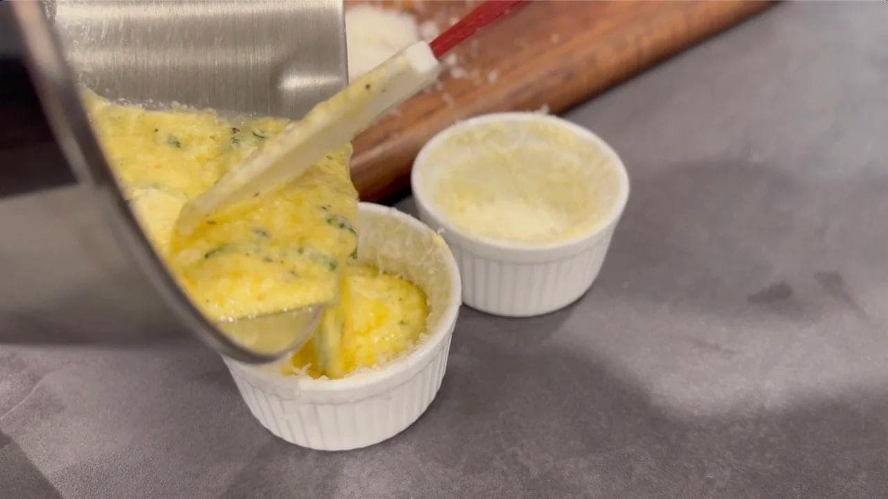 Pouring egg mixture into ramekins coated with melted butter and Parmesan cheese for baking.