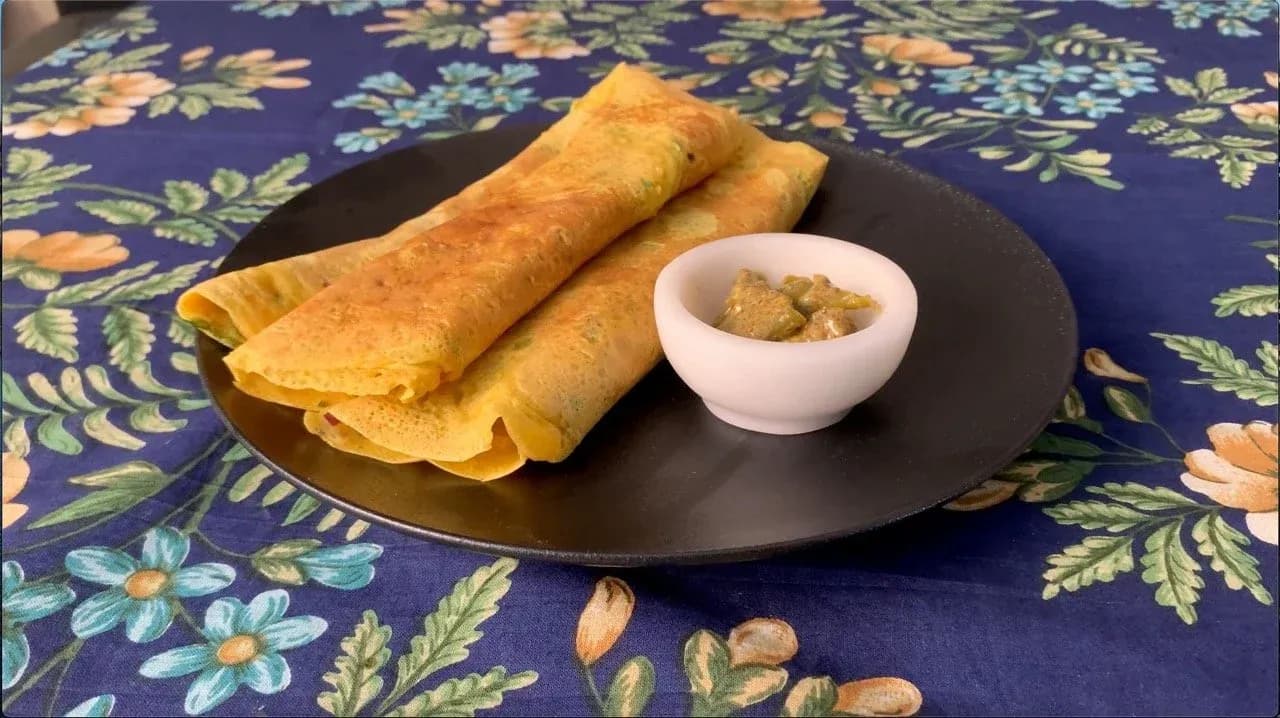 Two golden crispy besan chillas rolled cigar-style on a black plate with a white bowl of pickle chutney, on a blue floral tablecloth.