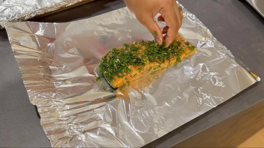 Lemon herb baked salmon fillet with green herb crust on aluminum foil, hand touching the cooked fish.