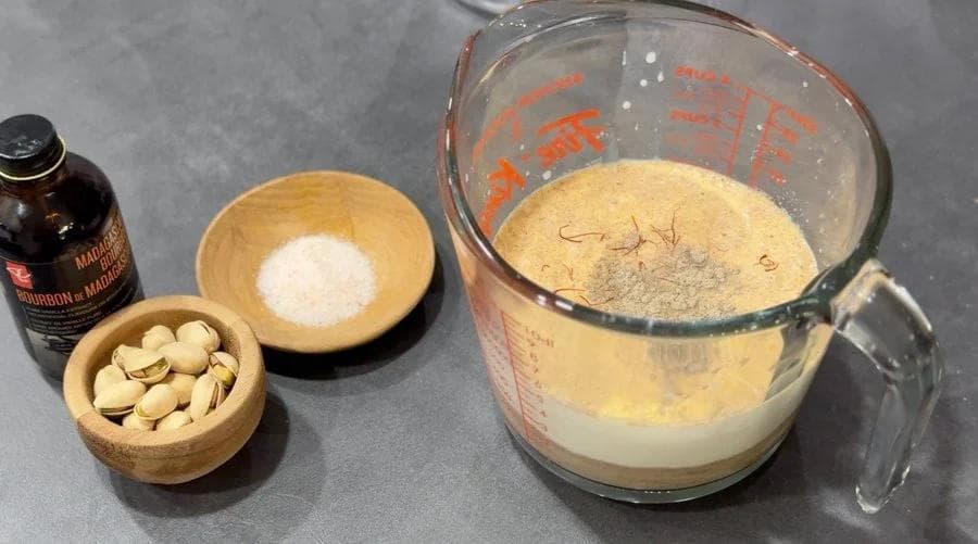 Ingredients for kheer ice cream: roasted semolina in measuring cup, condensed milk bottle, salt in wooden bowl, pistachios, and milk mixture in blender.
