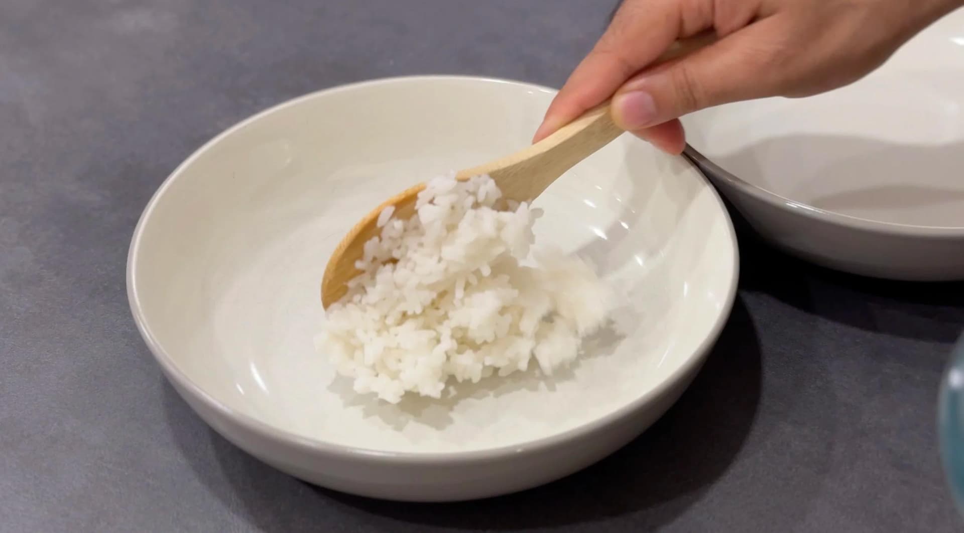 Hand spooning cooked white rice into a white bowl for assembling a poke bowl.