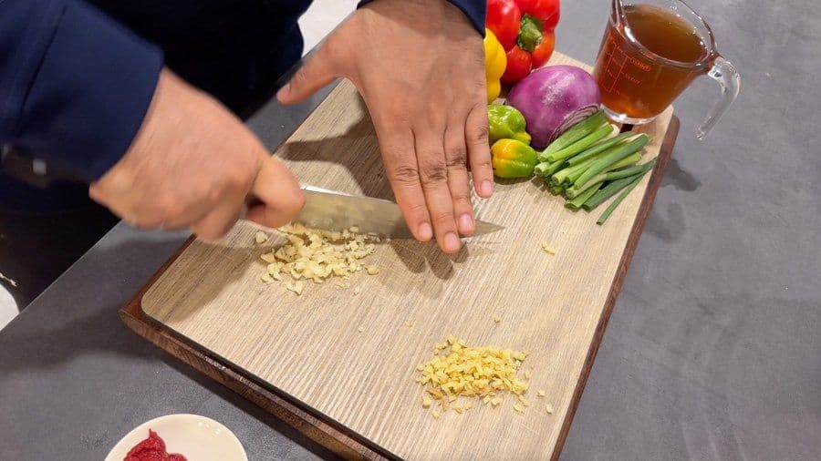 Chef mincing garlic on a wooden cutting board with fresh vegetables and ingredients nearby for Jamaican pepper steak.