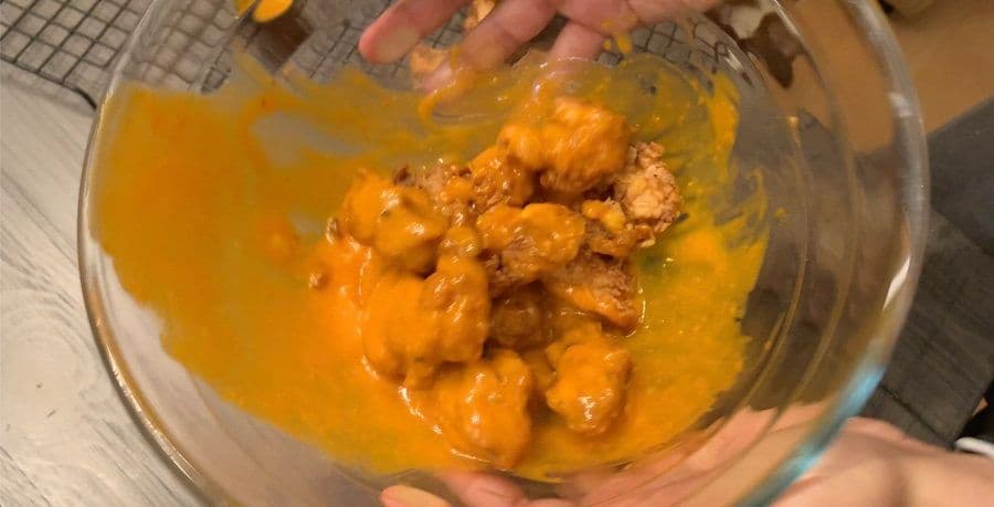 Crispy chicken pieces coated in orange buffalo sauce in a clear glass bowl being held by hands.