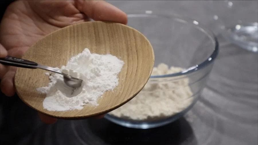 Hand holding wooden spoon with white protein powder over bowl of dry ingredients for pumpkin cookies.