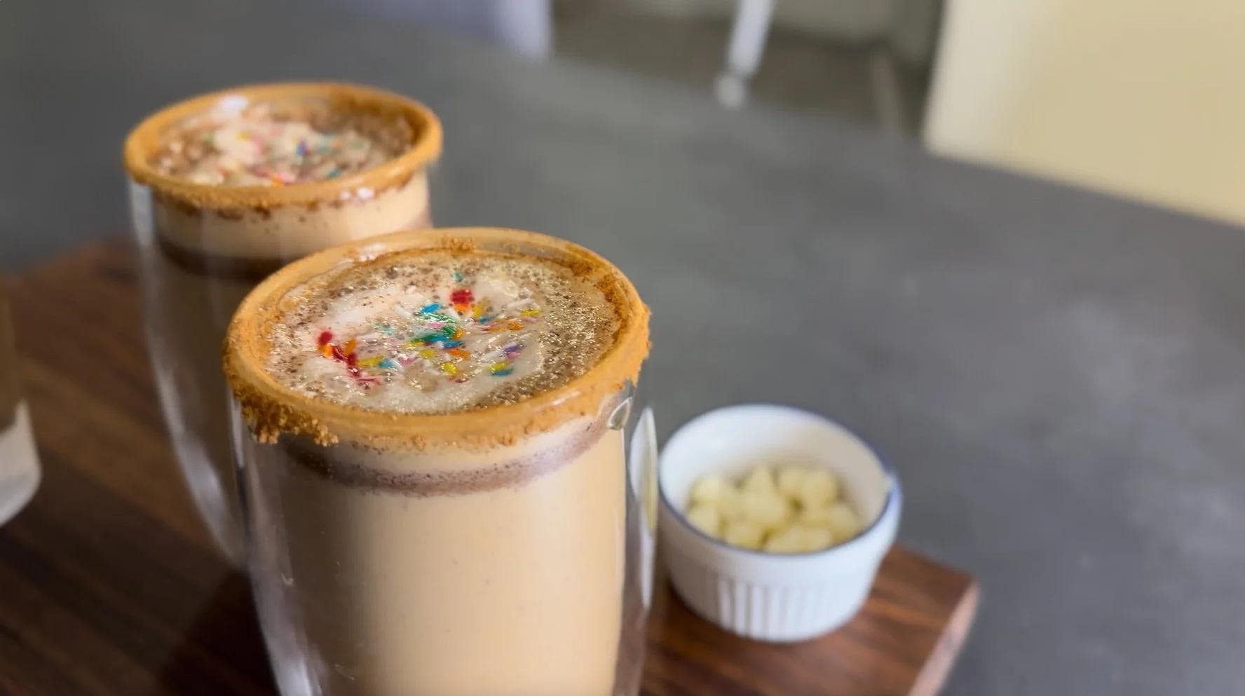 Two sugar cookie lattes with cookie crumble rim and colorful sprinkles, served with whipped cream on the side.