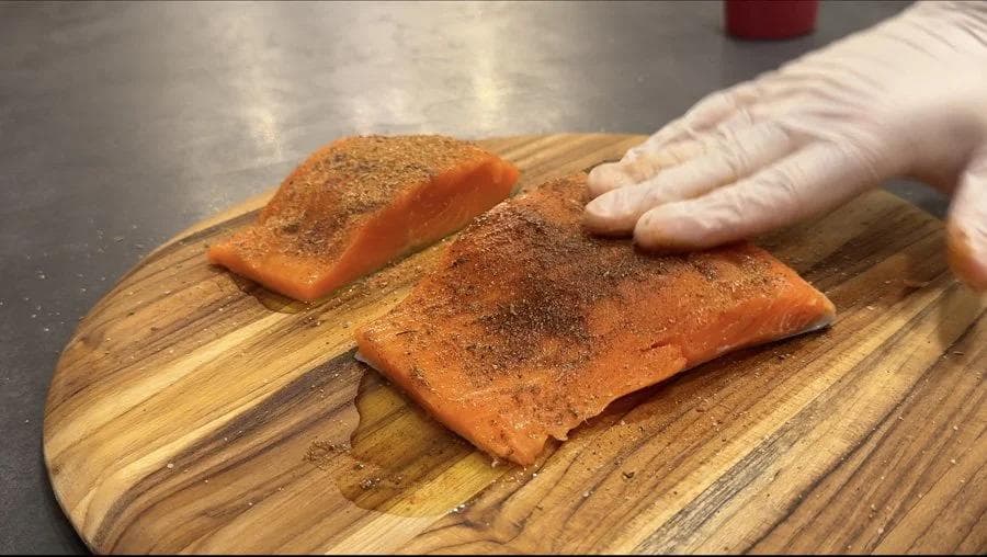 Two salmon fillets seasoned with blackening spice on a wooden cutting board, being rubbed with spices.