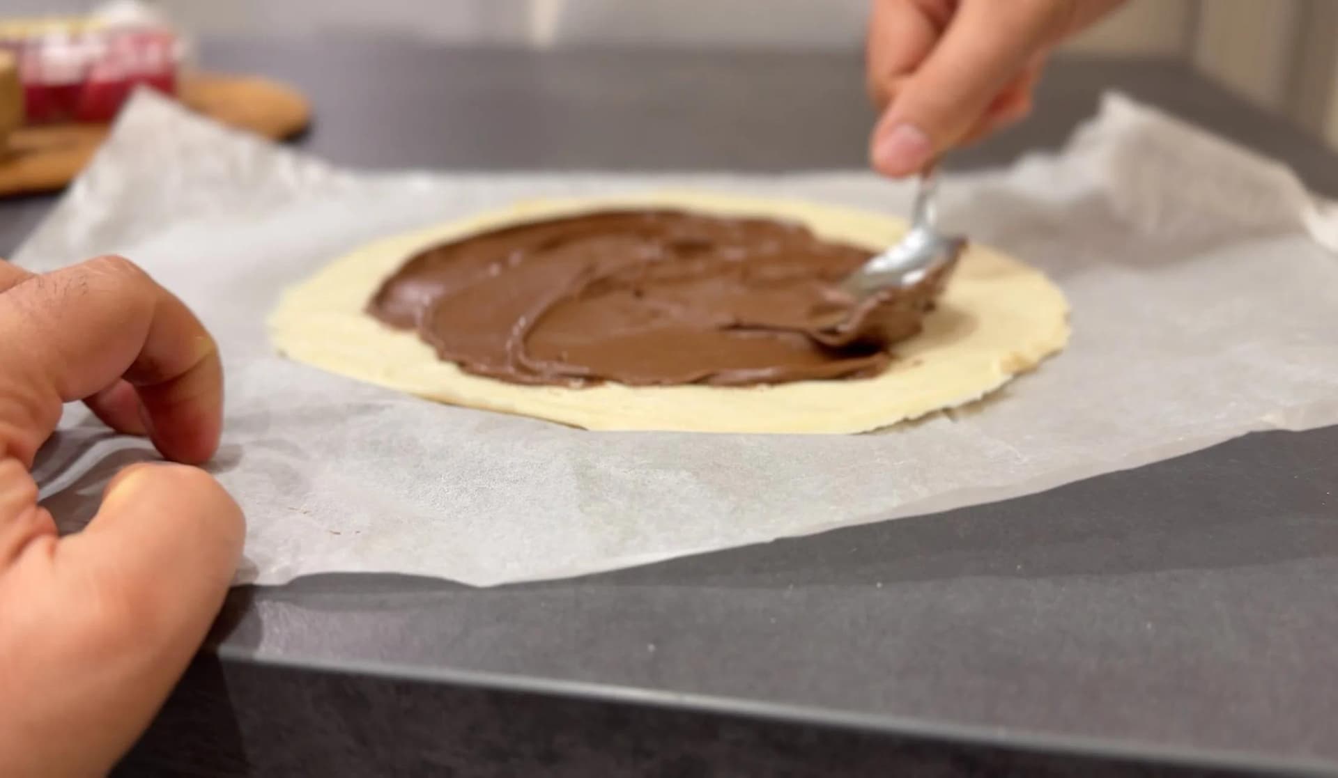 Spreading chocolate hazelnut spread on a puff pastry circle with a spoon, leaving a border around the edges.