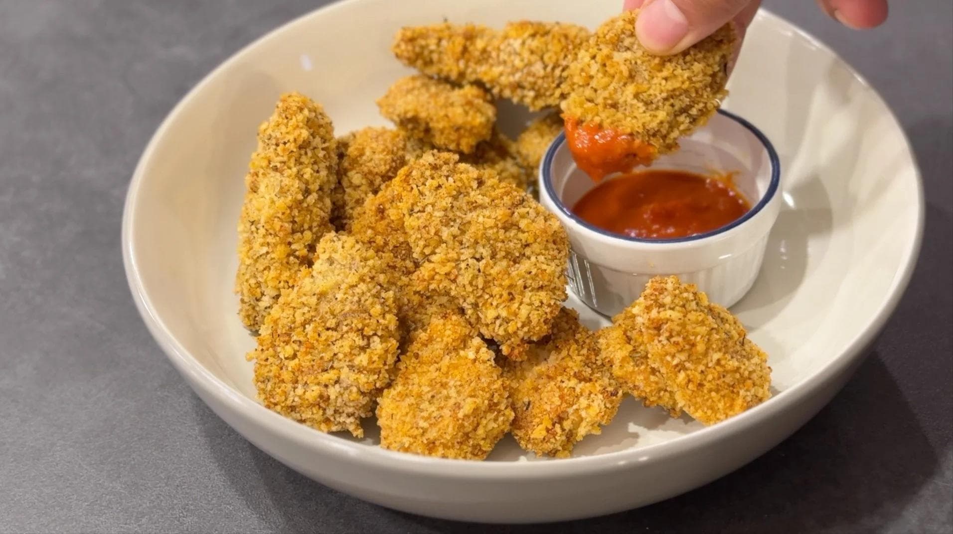 Crispy golden panko chicken nuggets on a white plate with a small bowl of red dipping sauce, one piece being dipped.