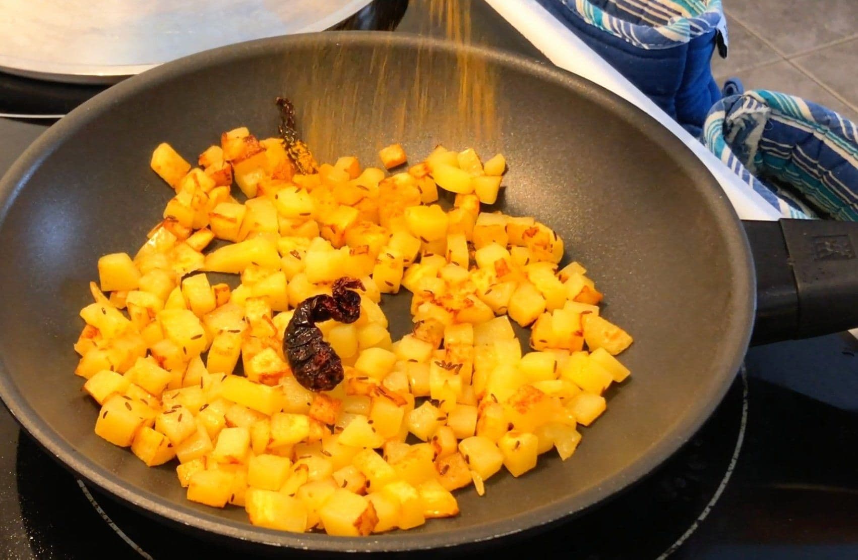 Spiced diced potatoes sautéed in a skillet with cumin seeds and a dried chili, being seasoned with turmeric powder.
