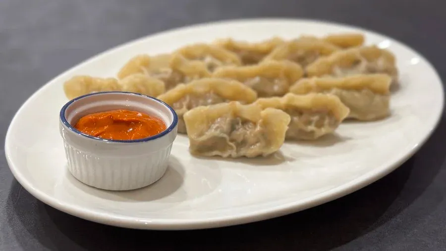 Steamed chicken momos on a white plate with a small bowl of orange dipping sauce