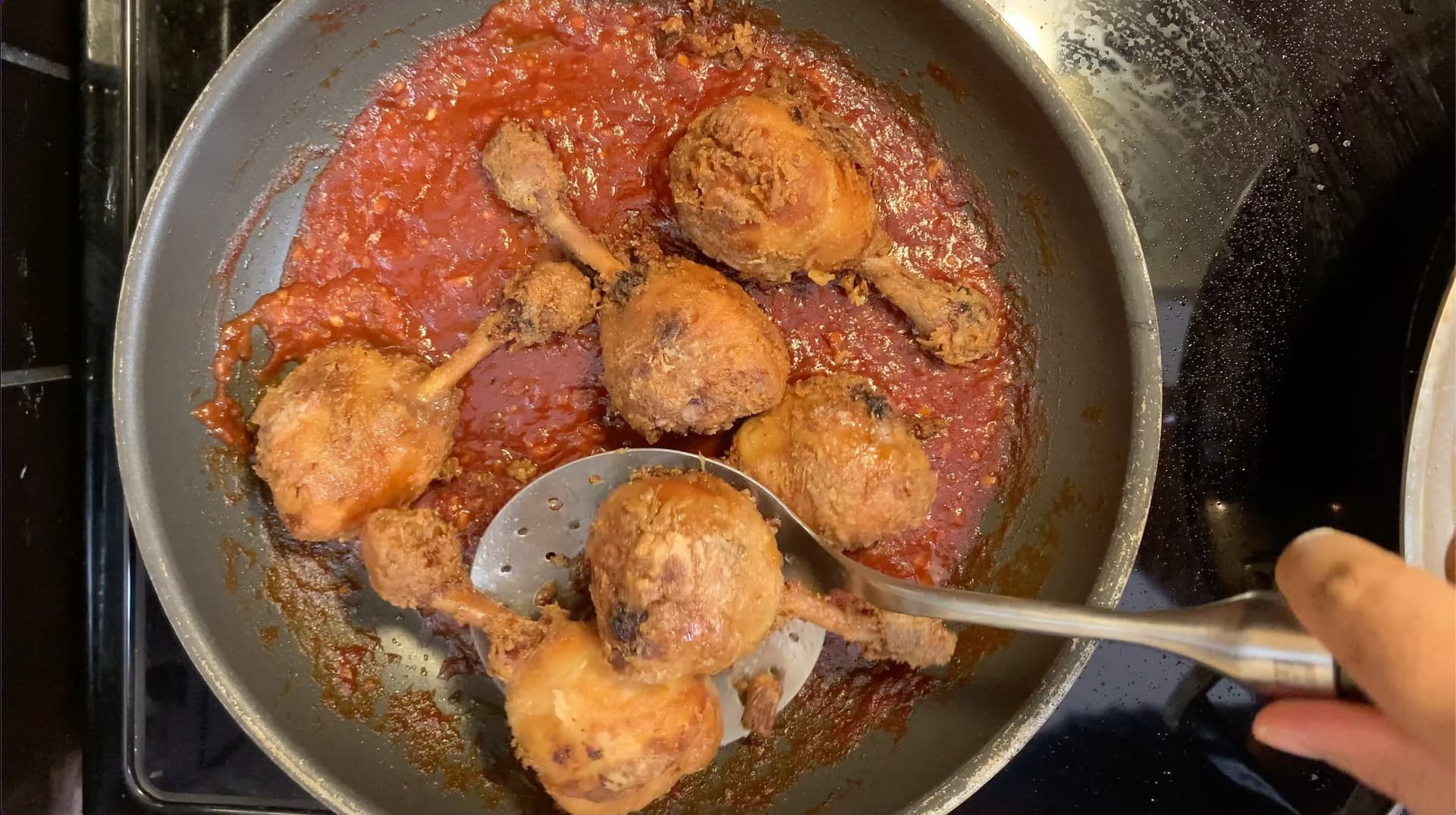 Golden fried chicken drumsticks coated in red spiced sauce in a pan with a slotted spoon.