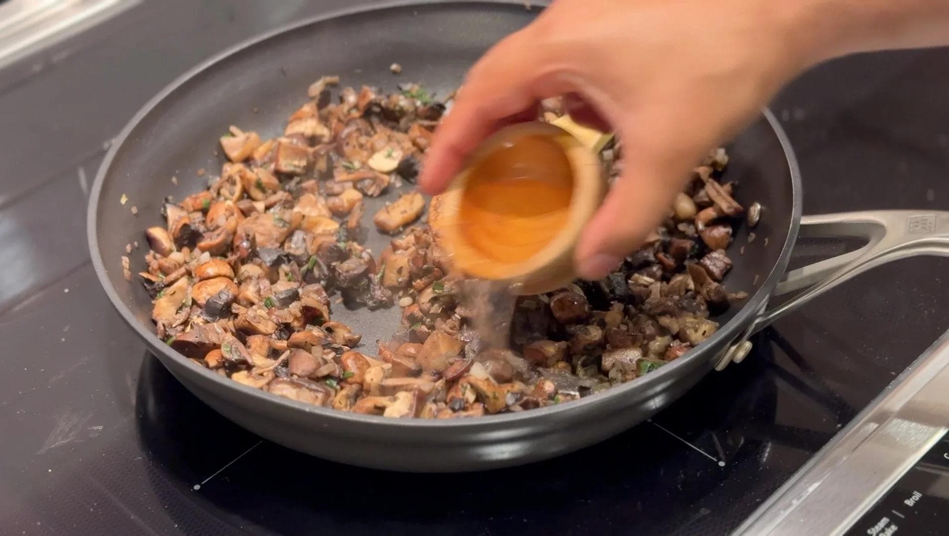 Hand pouring beaten egg into a skillet with sautéed mushrooms and shallots on stovetop.