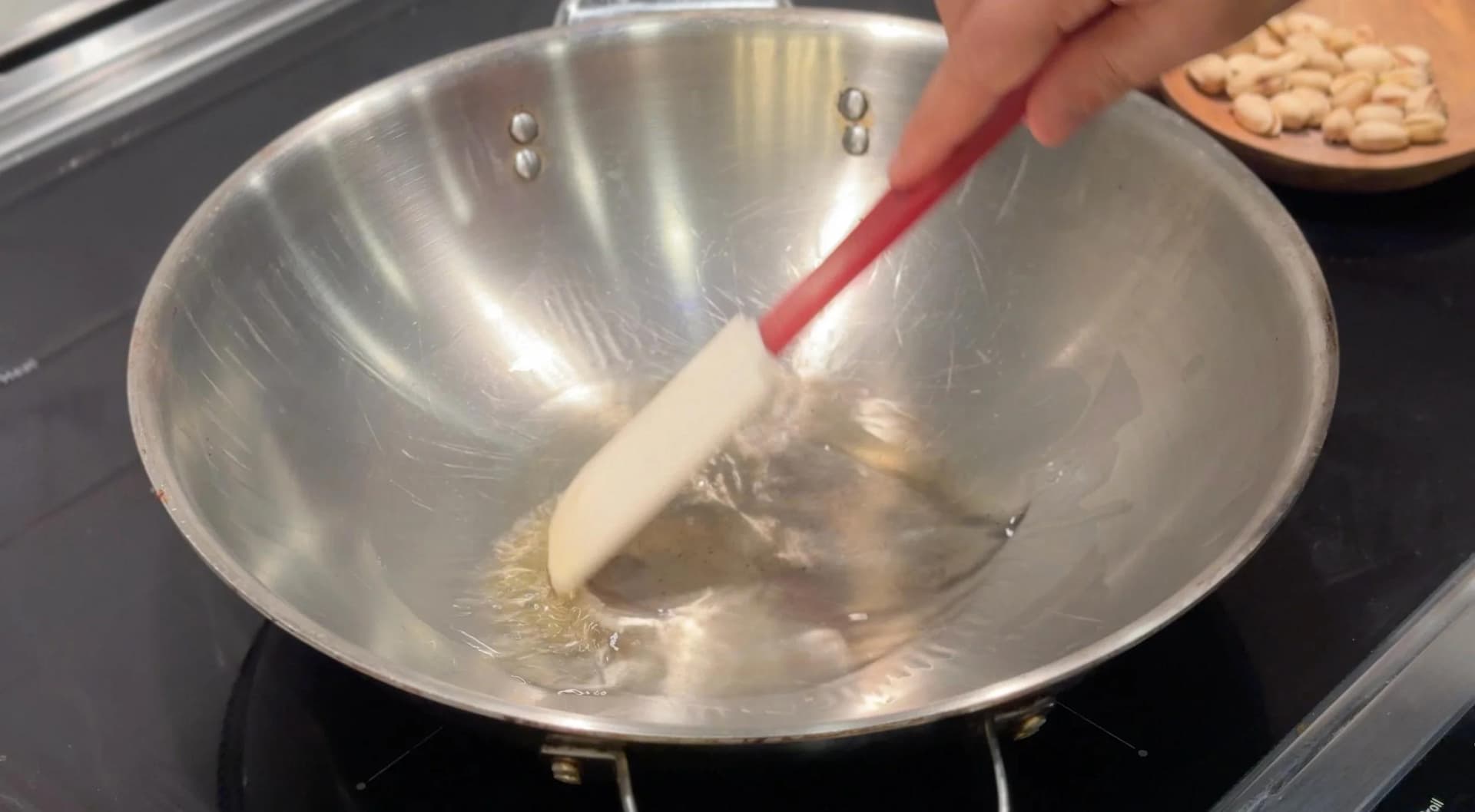 Stirring roasted semolina in a wok with a spatula, with nuts visible in a bowl nearby.