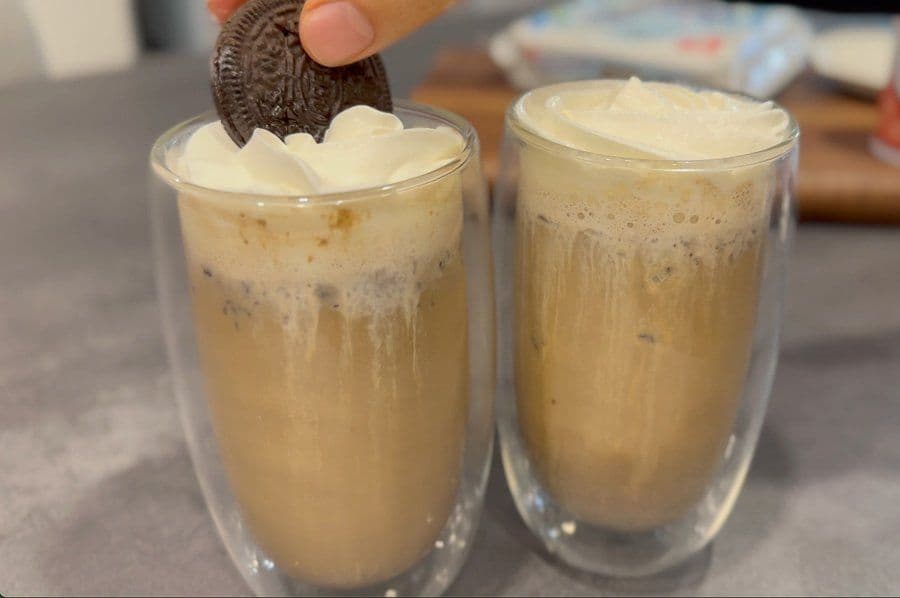 Cookies and cream Oreo lattes topped with whipped cream and an Oreo cookie being added to the left glass.