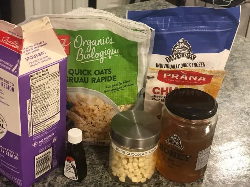 Ingredients for blueberry oatmeal displayed on countertop: oats, honey, white chocolate chips, chia seeds, and frozen blueberries.