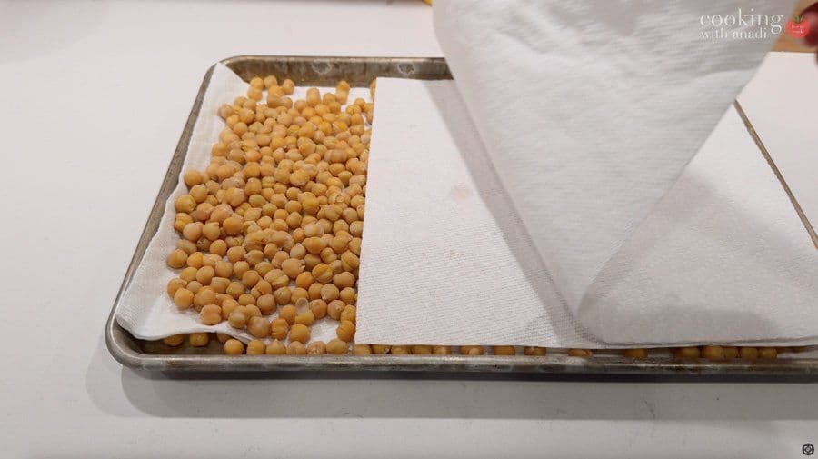 Chickpeas spread on parchment paper in a baking tray being patted dry with paper towels.
