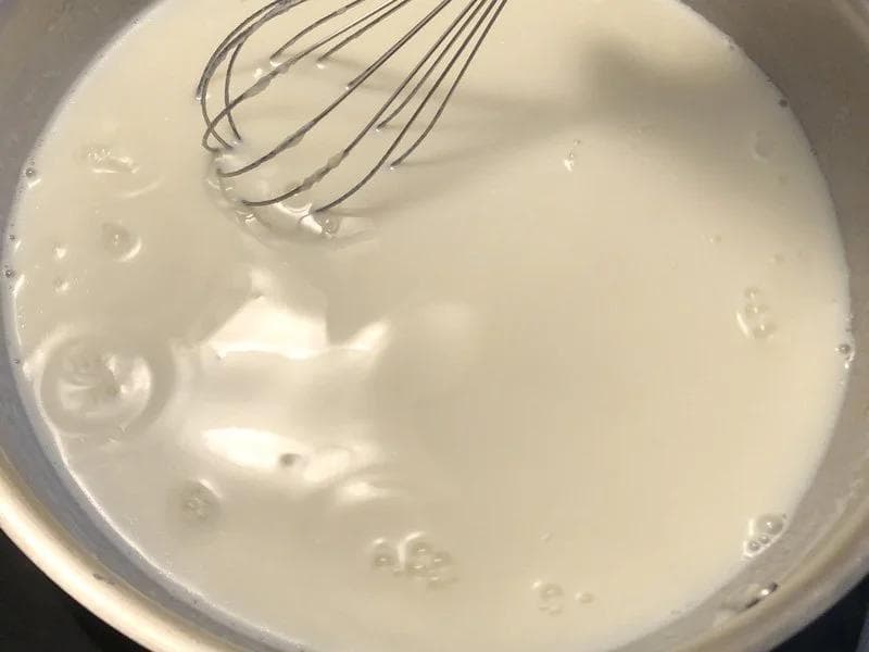 Creamy white béchamel sauce in a pot with a whisk, showing smooth texture after milk is incorporated.