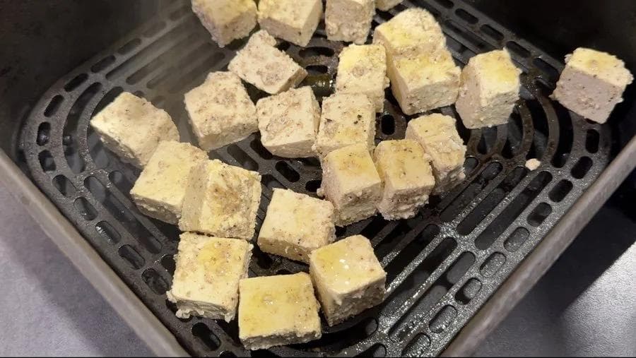 Tofu cubes lightly coated in oil arranged in a single layer in an air fryer basket before cooking.