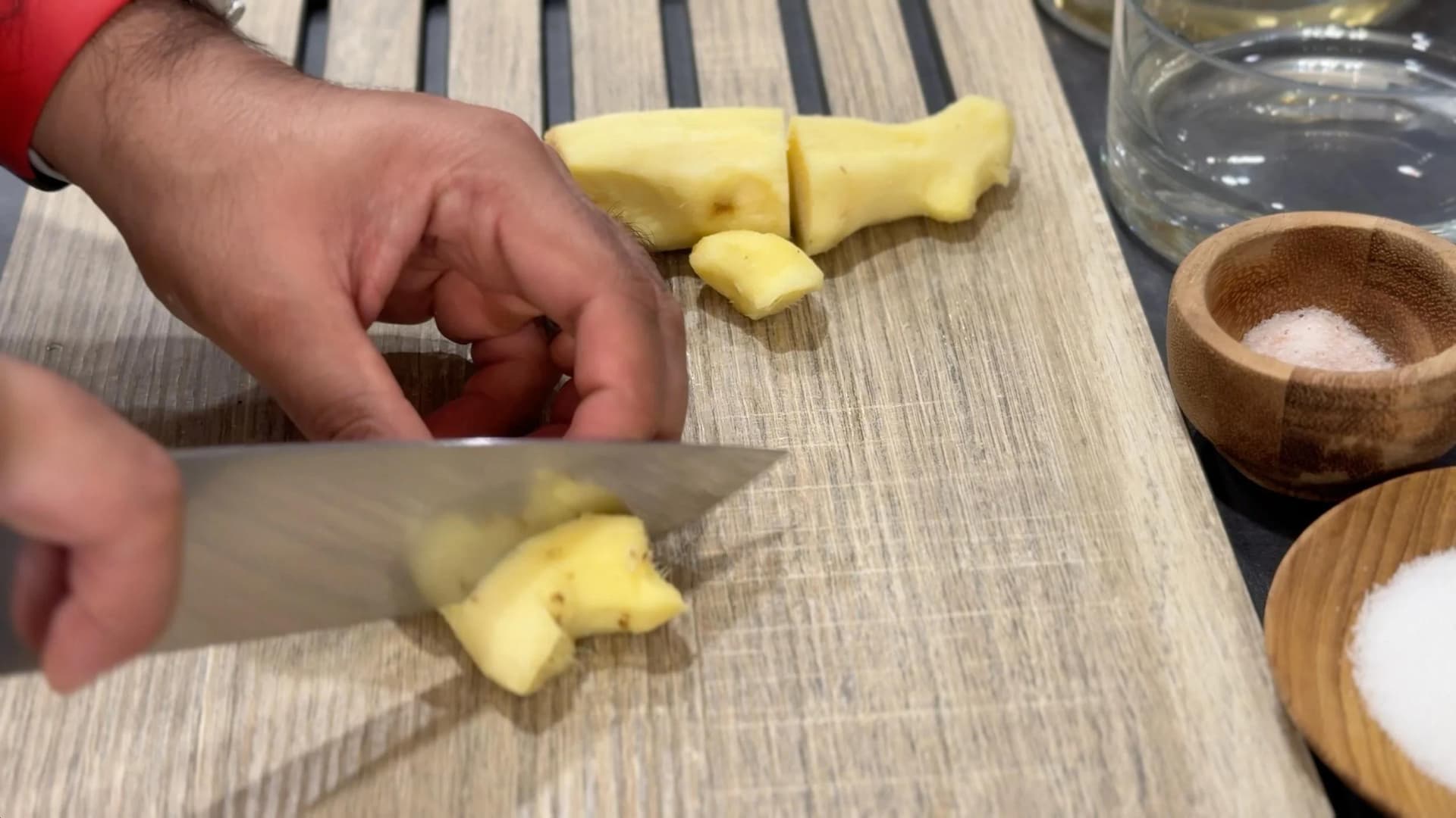 Hand slicing thin pieces of peeled ginger on a wooden cutting board with a knife.